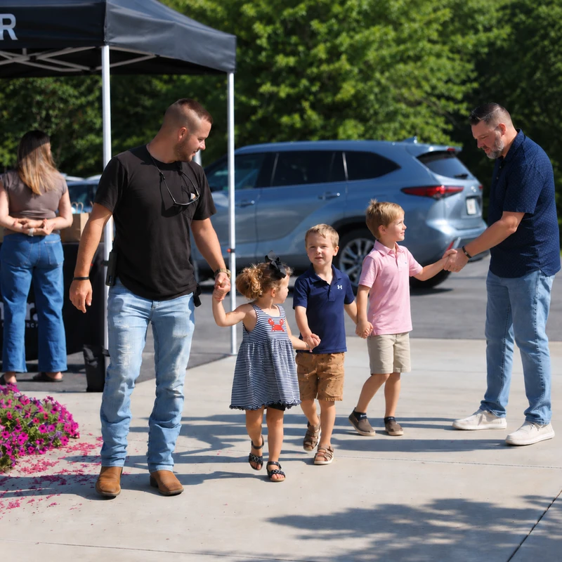 A man leads a little girl and boy by the hand while another man shakes hands with a second boy as they arrive at an outdoor event on a sunny day.