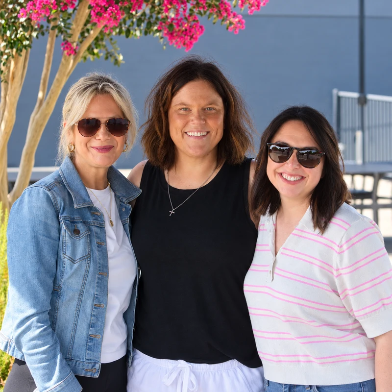A group portrait of three smiling women posing together outdoors under a tree with vibrant pink flowers. They are dressed in casual summer clothes.