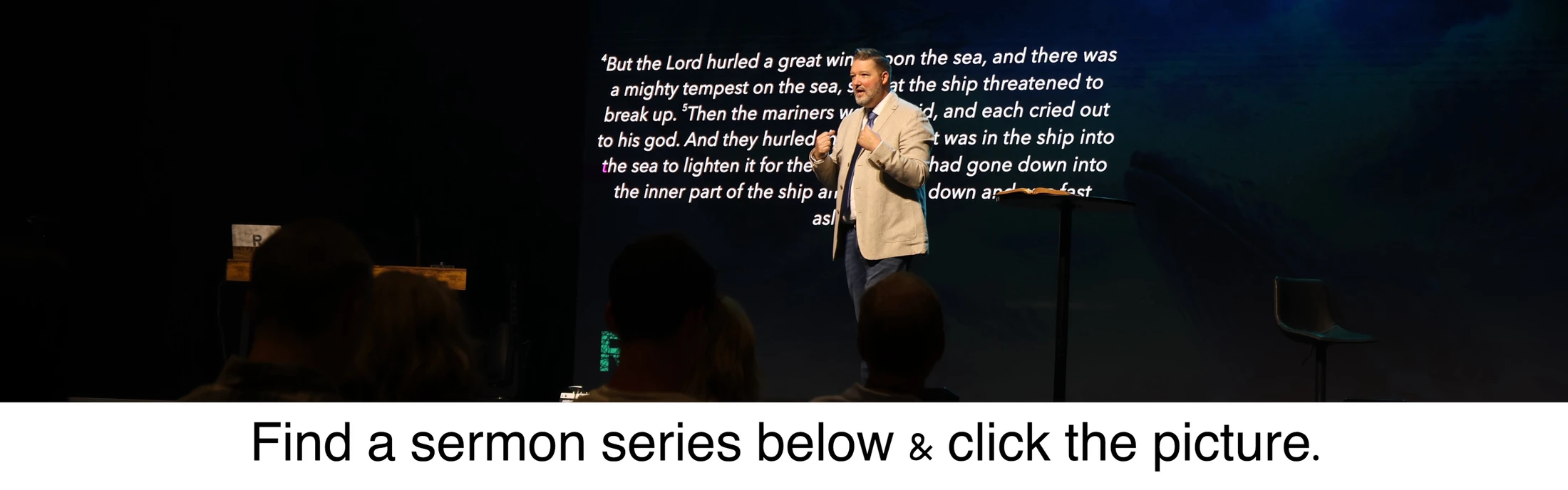A pastor in a tan jacket preaches on a dark stage. Behind him, a large screen displays a Bible verse. The audience is seen from the back.