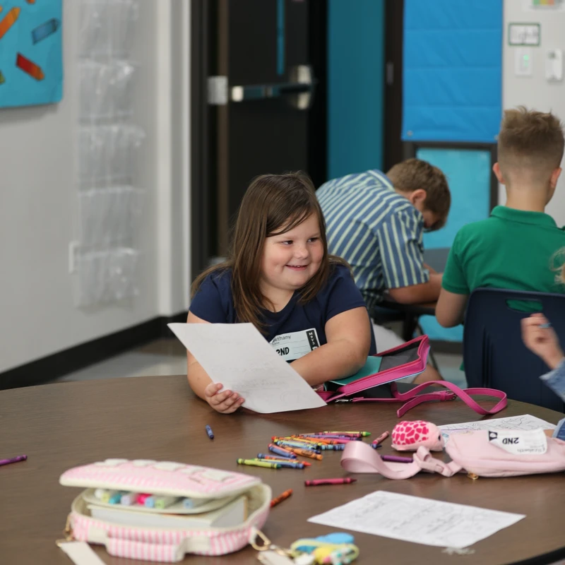 A young girl with brown hair sits at a round table in a classroom, smiling as she holds a piece of paper. Crayons and a pencil case are on the table.