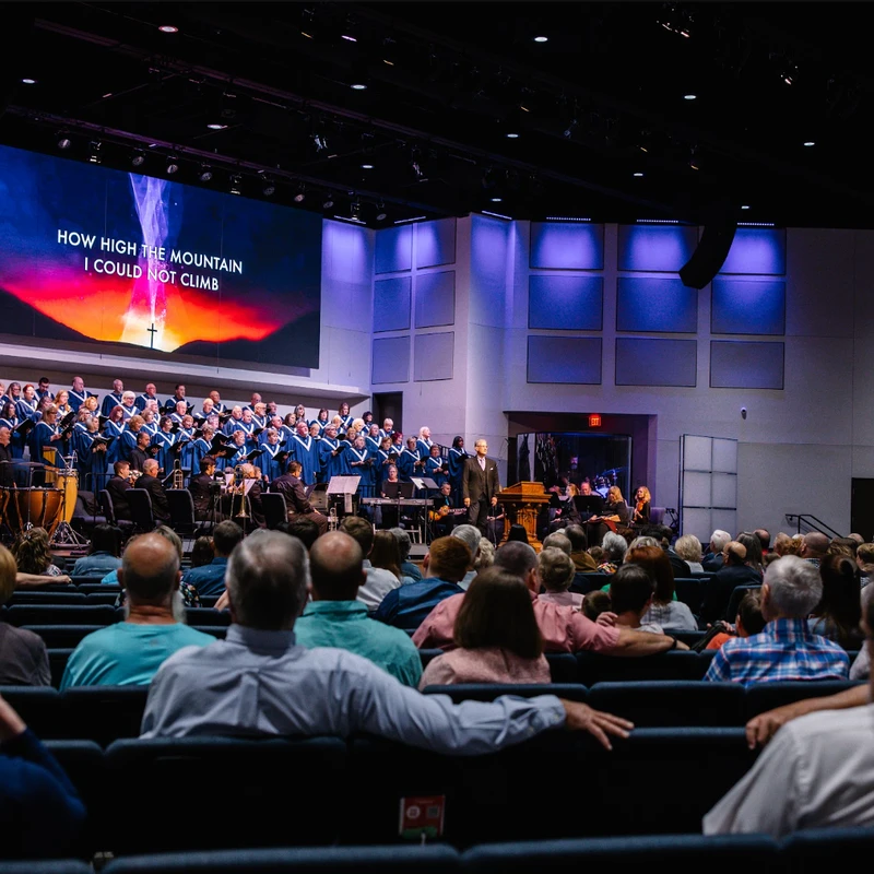 View from the audience of a large choir in blue robes and an orchestra performing on stage in a modern church. A screen behind them displays song lyrics.