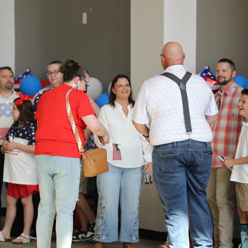 A group of adults and children gather indoors for a patriotic celebration, decorated with red and blue balloons and American flags, smiling and socializing.
