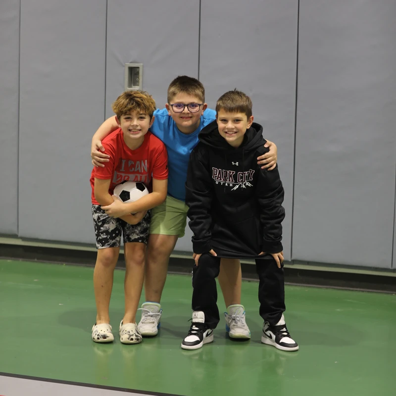 Three young boys smile as they stand with their arms around each other on a green gym floor. The boy on the left holds a soccer ball.