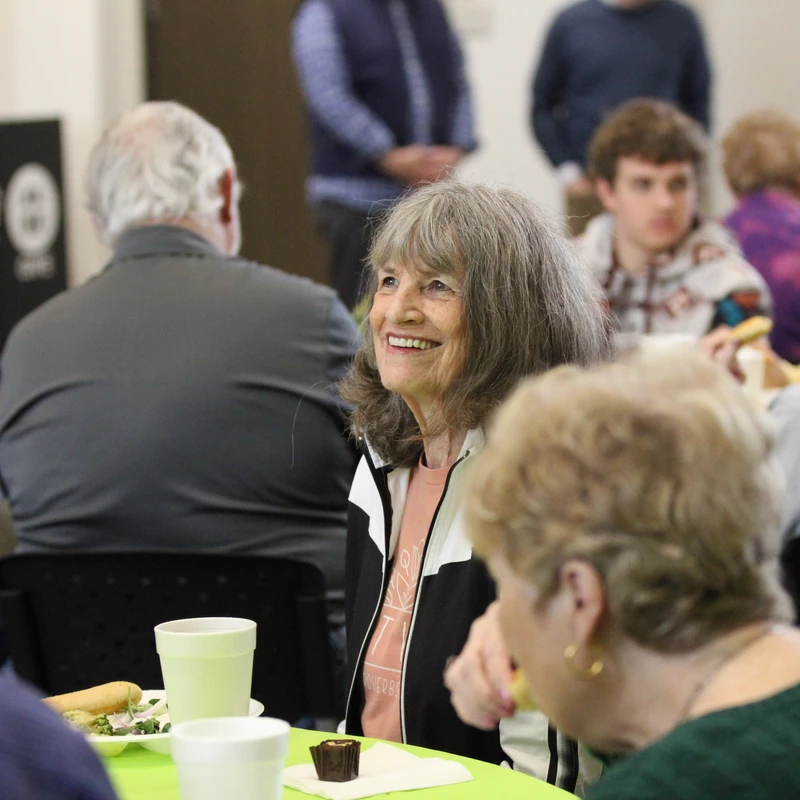 An older woman with shoulder-length gray hair smiles warmly while seated at a table during a crowded community luncheon.