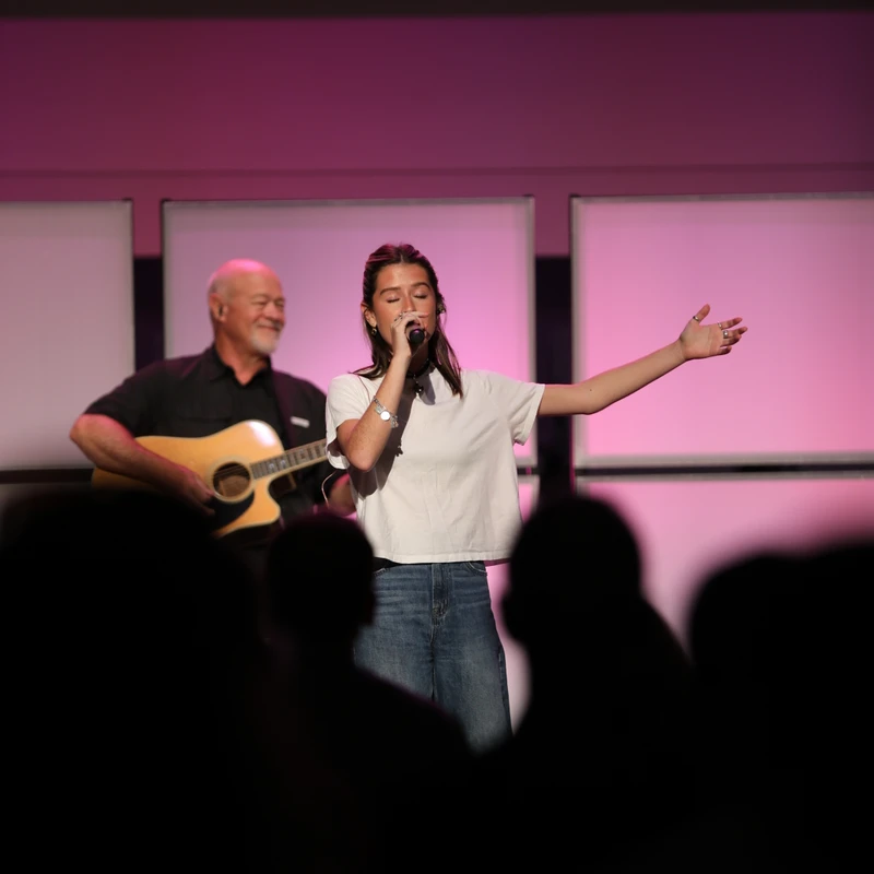 A young woman sings into a microphone on a pink-lit stage, with a man playing guitar behind her. The audience watches from the foreground as silhouettes.