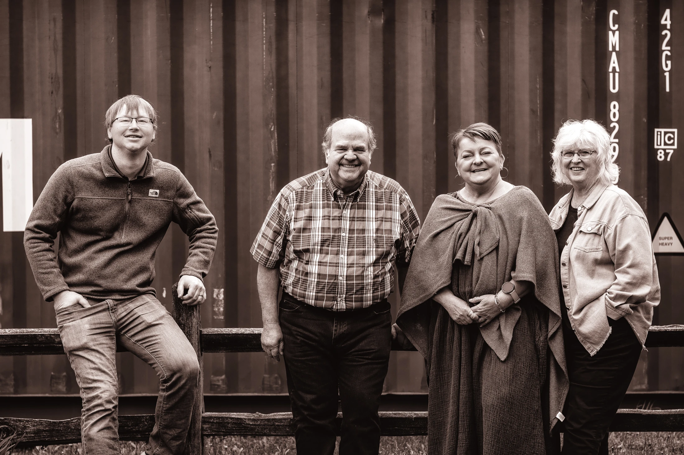 A sepia-toned group photo of four smiling adults, two men and two women, posing casually in front of a dark, corrugated metal shipping container.