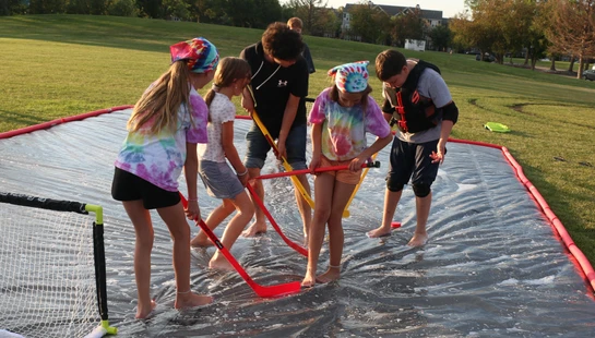 A group of children and teens play a game of hockey on a wet, soapy slip-n-slide set up on the grass in a park.