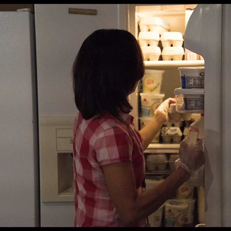 A woman with dark hair wearing a plaid shirt and disposable gloves reaches into a brightly lit, well-stocked refrigerator filled with egg cartons and yogurt.
