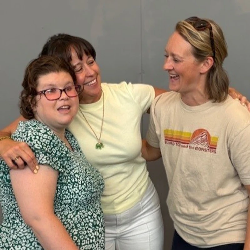 Three women stand close together, smiling and laughing. The woman in the center has her arms around the other two in a happy, friendly embrace.