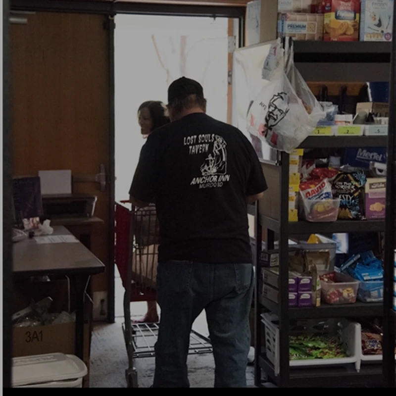 A man and a woman push a shopping cart through a small store or food pantry. The man's back is to the camera, and shelves of food are on the right.