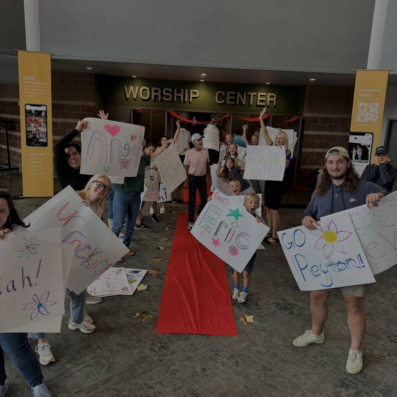 A group of smiling people of all ages hold up handmade signs to welcome others along a red carpet leading to a Worship Center.