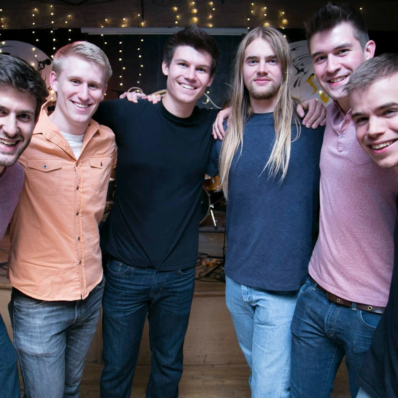 A group of six young men stand with arms around each other, smiling for the camera. They are in a dimly lit room with string lights in the background.