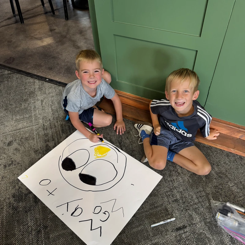 Two young boys sit on a grey carpeted floor, smiling up at the camera. Between them is a large drawing of a face they have made with markers.