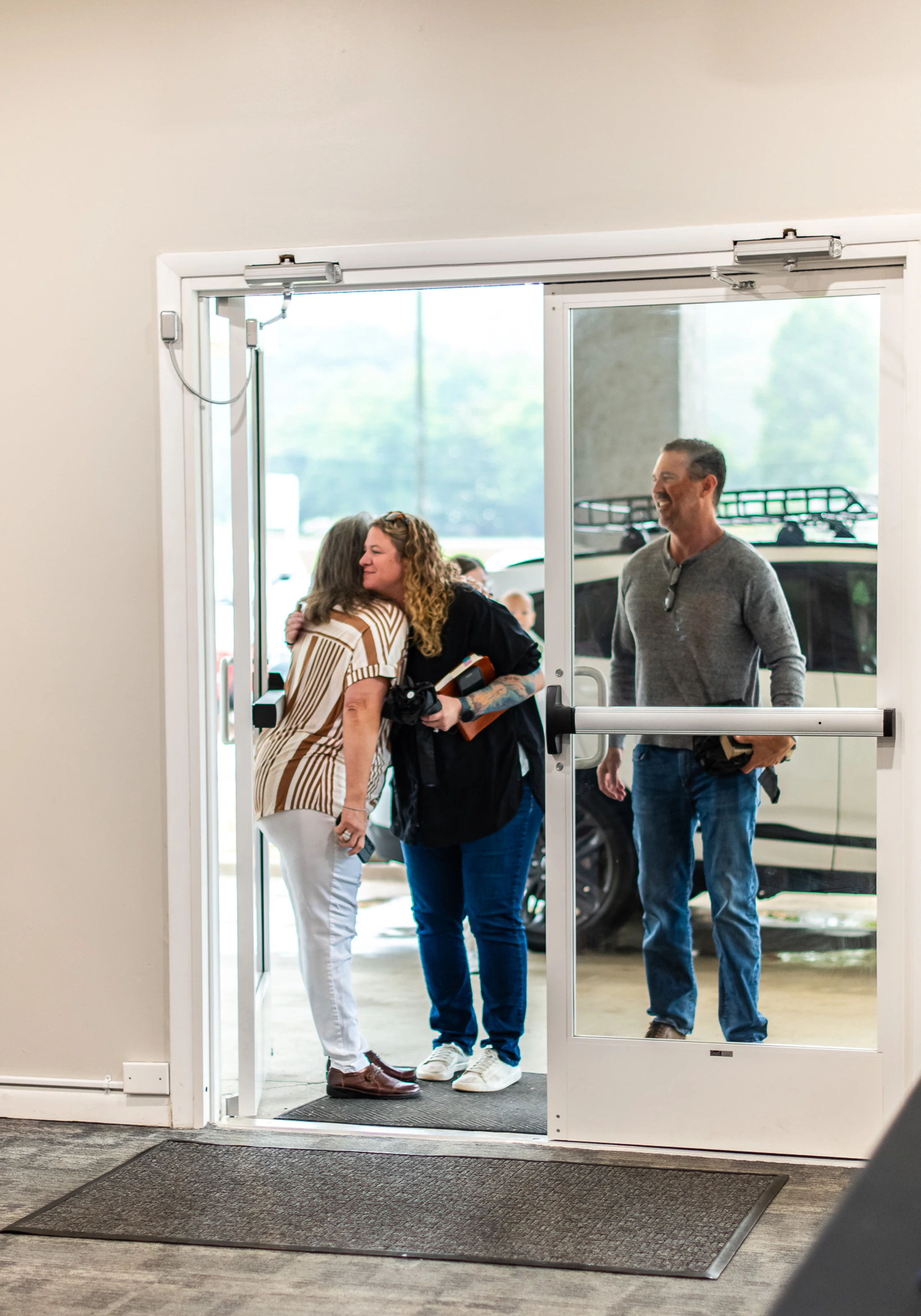 Two women embrace in a hug in an open glass doorway while a man stands smiling nearby, with a white car visible outside.
