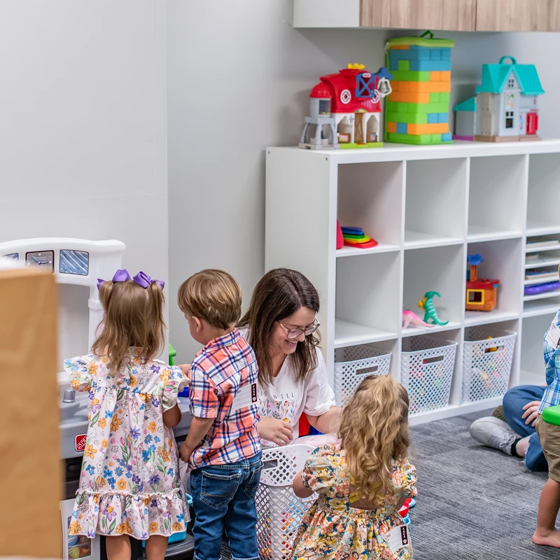 A female teacher sits on the floor with three young children in a classroom. They are gathered around a toy kitchen and other toys in a play area.
