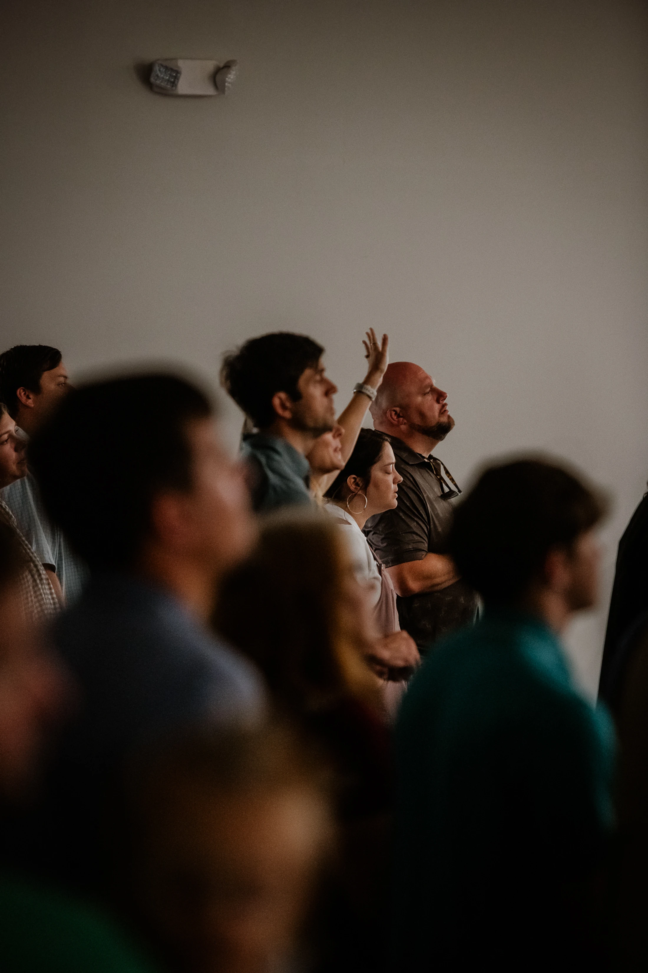 A diverse group of people stand together looking upwards. One woman has her hand raised, and a man next to her stands with arms crossed, looking on intently.