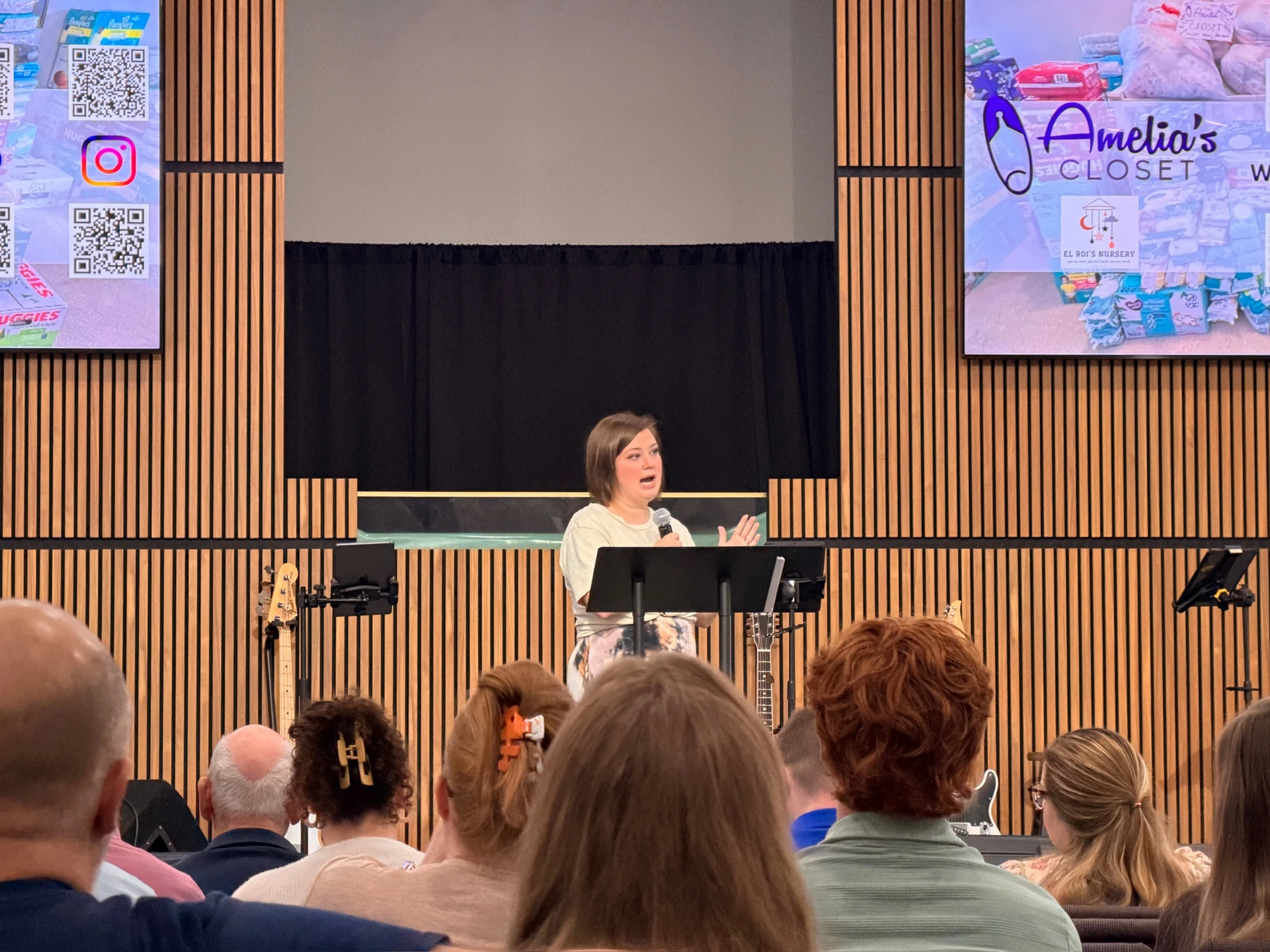 A woman stands at a podium on a stage with a modern wood-slat wall, speaking to an audience. Screens on either side show a charity logo for "Amelia's Closet".