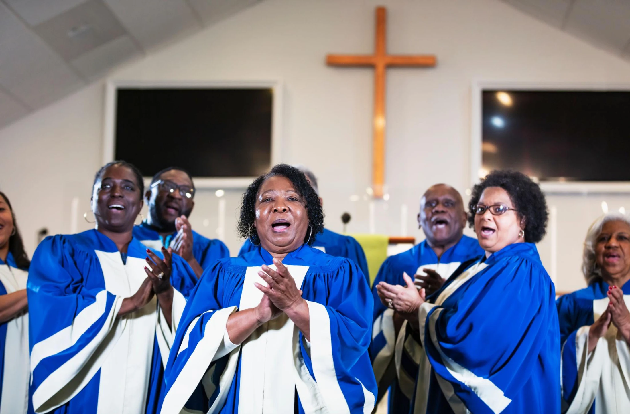 A group of African American men and women in blue and white robes sing joyfully as a gospel choir in a church, with a wooden cross in the background.