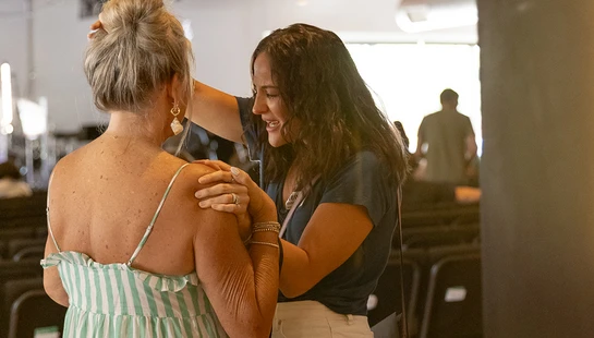A smiling woman with a 'Prayer Team' lanyard gently places her hands on another woman's shoulder and head in a comforting gesture in a large hall.