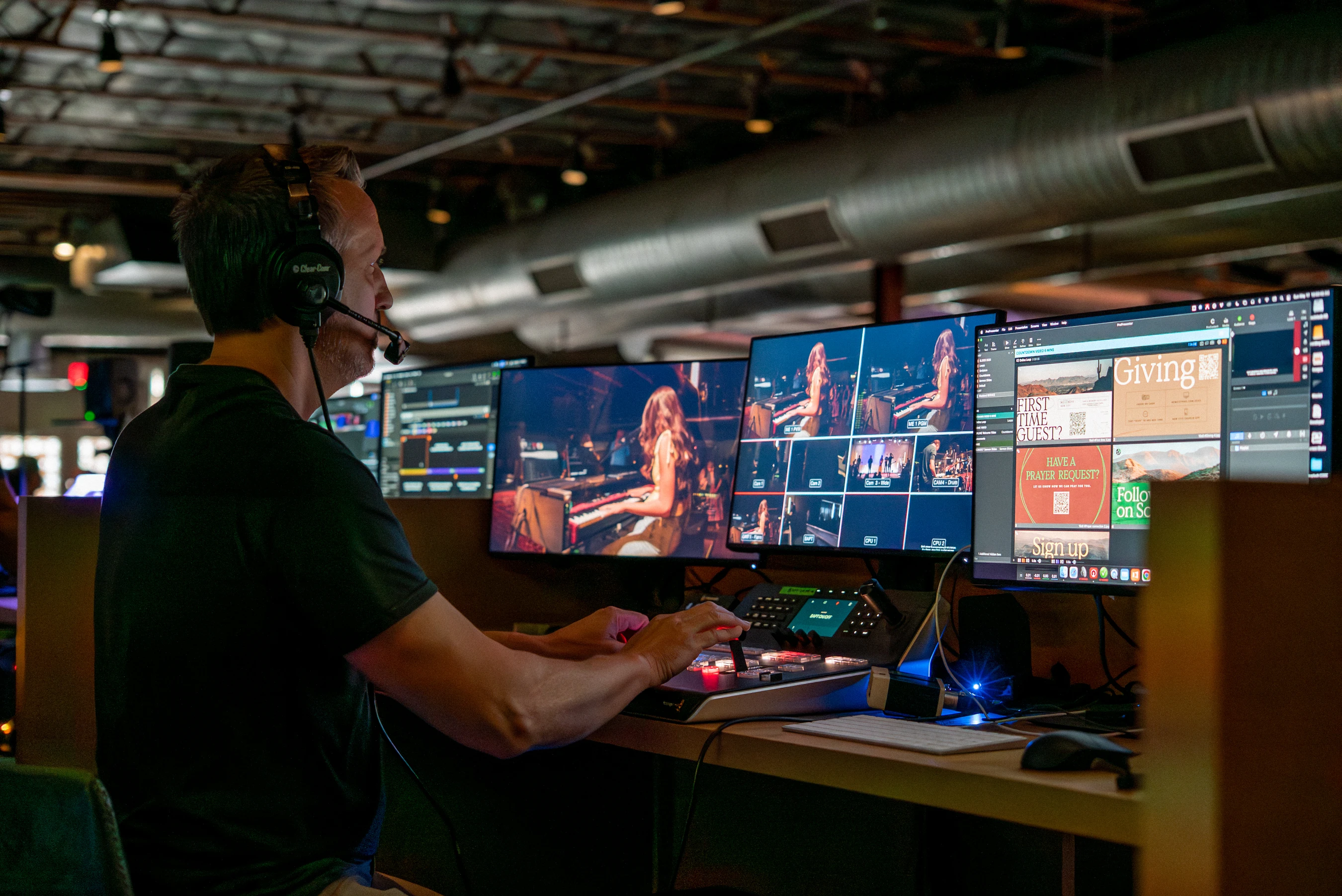 A man wearing a headset sits at a control desk with multiple monitors, directing a live video broadcast of a church service from a production booth.