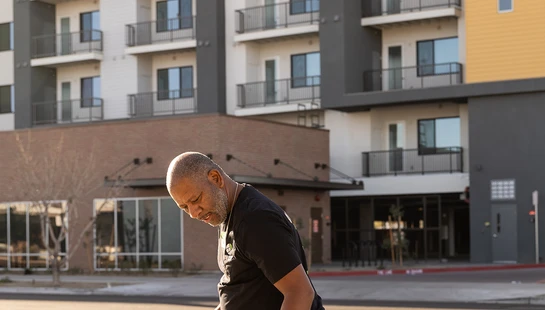 A man wearing blue gloves holds a white trash bag while picking up litter in a parking lot, with a modern apartment building in the background.