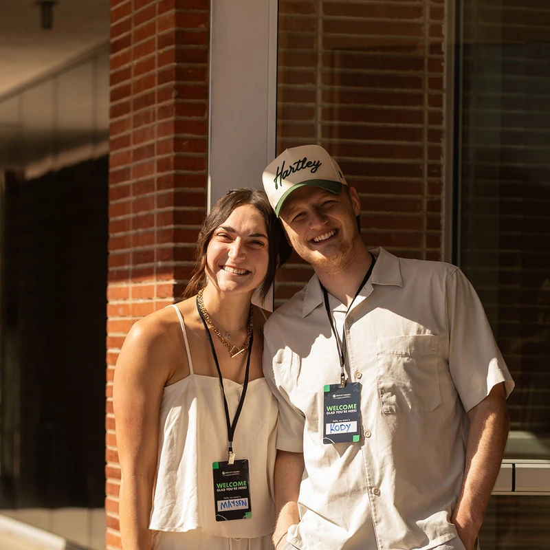 A young man and woman smile warmly at the camera. They are standing outside a brick building and both wear lanyards with name tags that say 'Welcome'.
