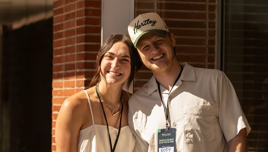 A young man and woman smile warmly at the camera. They are standing outside a brick building and both wear lanyards with name tags that say 'Welcome'.