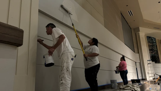 Three painters apply a fresh coat of light paint to a large interior wall. A woman uses a roller on a long pole while a man uses a brush for details.