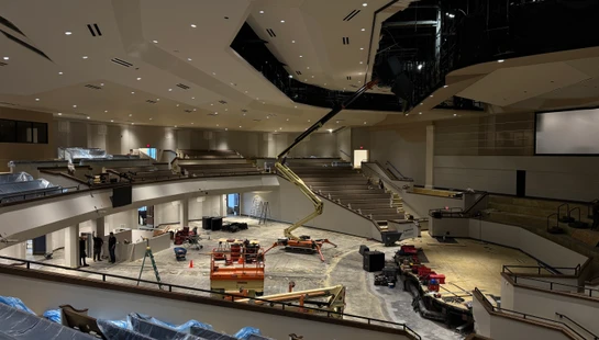 A wide view of a large auditorium during renovation, with a boom lift in the center and tiered seating covered in plastic sheeting.