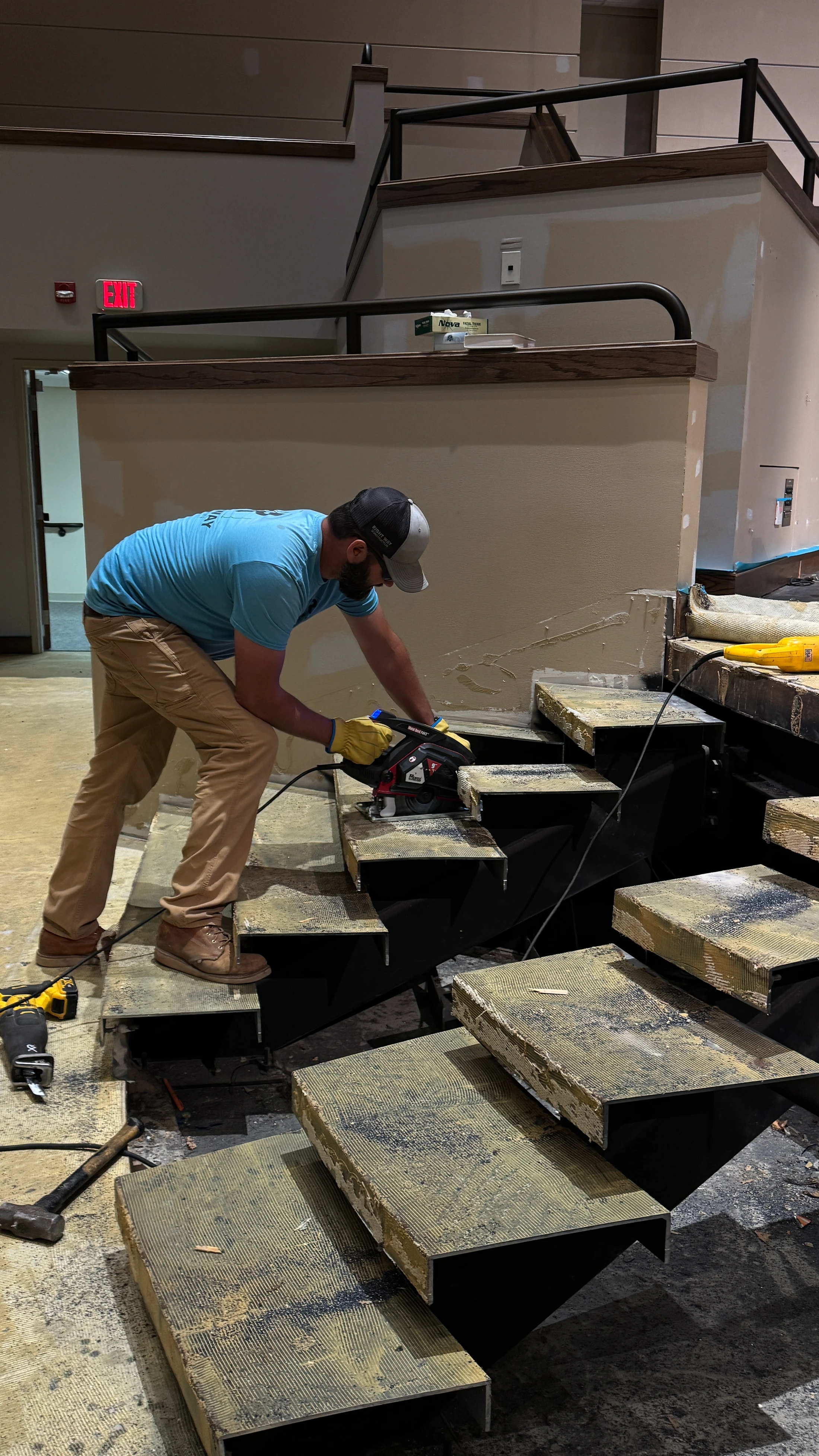 A man wearing work clothes and gloves uses a circular saw on a set of stairs that are under construction inside a building.