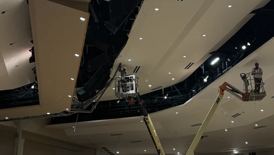 Two construction workers on hydraulic lifts work on the high, partially demolished ceiling of a large auditorium during a major renovation project.