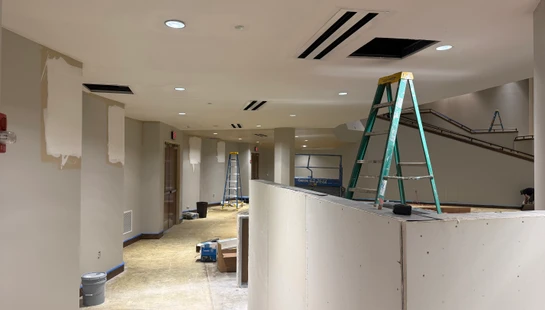 A long hallway in a commercial building is under construction, with ladders, unfinished drywall, a scissor lift, and exposed floors.