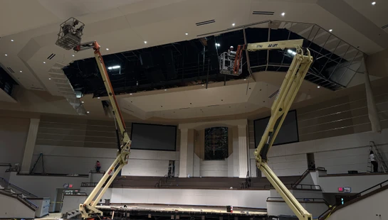 Two large boom lifts with workers reach up to the high, partially deconstructed ceiling of an auditorium undergoing extensive renovation.