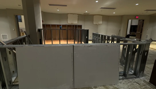 An office or large room undergoing renovation, with metal stud framing for new walls in the foreground and unfinished, patched walls in the background.