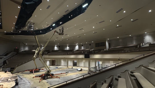 A wide-angle view of a large auditorium under renovation, with two boom lifts on the floor extending up to workers installing ceiling fixtures.