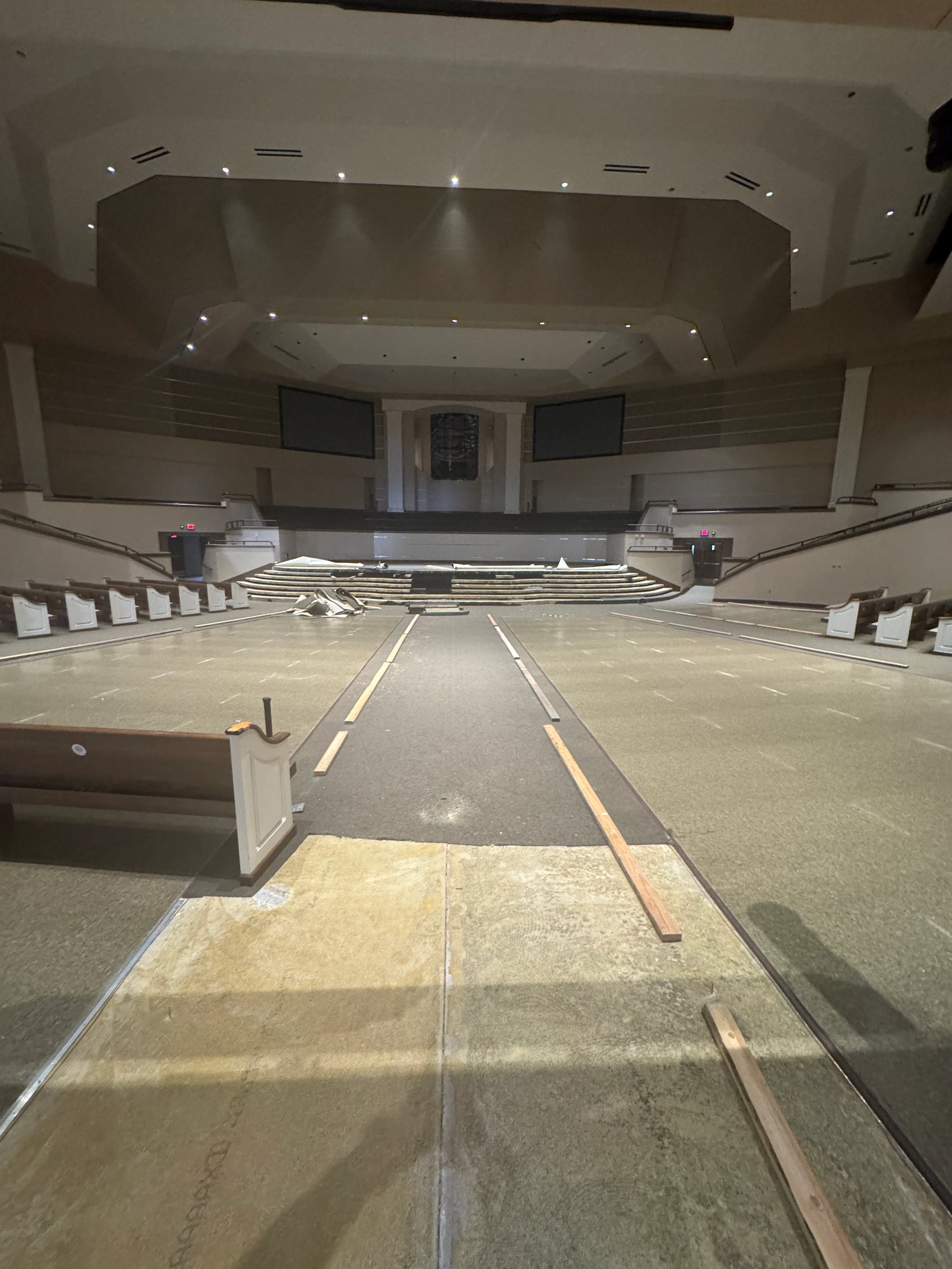 A wide shot of a large, empty church sanctuary undergoing renovation. Most pews are removed, leaving bare floors, with construction debris on the tiered stage.