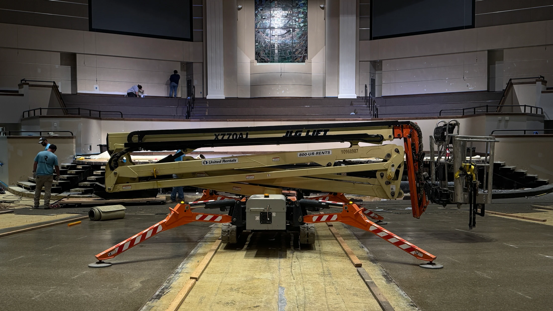 A JLG boom lift with orange spider-like legs sits in the center of a large auditorium during a renovation, with workers on the stage in the background.