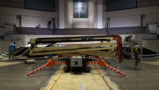 A JLG boom lift with orange spider-like legs sits in the center of a large auditorium during a renovation, with workers on the stage in the background.