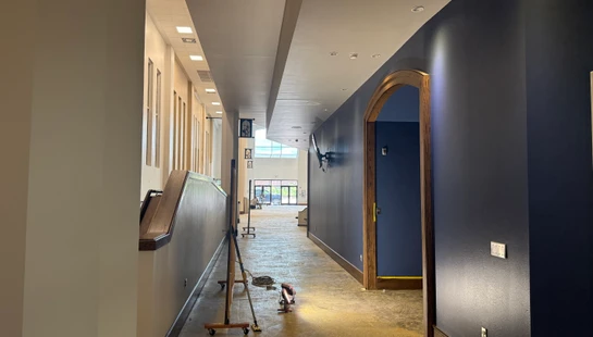 A long building hallway during renovation, with a dark blue wall opposite a light-colored wall, an unfinished concrete floor, and an arched wooden doorway.