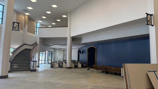 A spacious building lobby with a high ceiling undergoing renovation. The floor is bare concrete, pews are pushed aside, and a curved staircase is on the left.