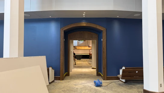 A room under construction with new blue paint on the walls, a wooden arched doorway, and construction materials like drywall and an industrial fan on the floor.