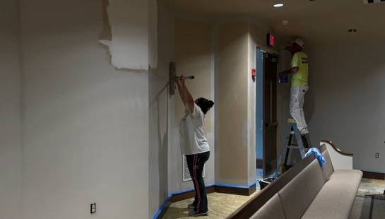 Two painters work in a hallway. A woman uses a pole sander on a wall, while a man on a stepladder paints above a doorway with a red exit sign.
