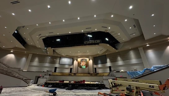A large church sanctuary during renovations, with a construction lift, bare floors, a deconstructed stage, and an open ceiling showing internal structures.