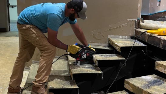 A construction worker in a blue shirt and cap uses a circular saw on a metal staircase during a renovation project, with tools scattered around.