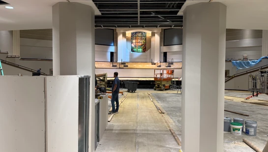 A large church sanctuary during renovation, seen from between two pillars. A worker, construction lift, and stained-glass window are visible in the background.