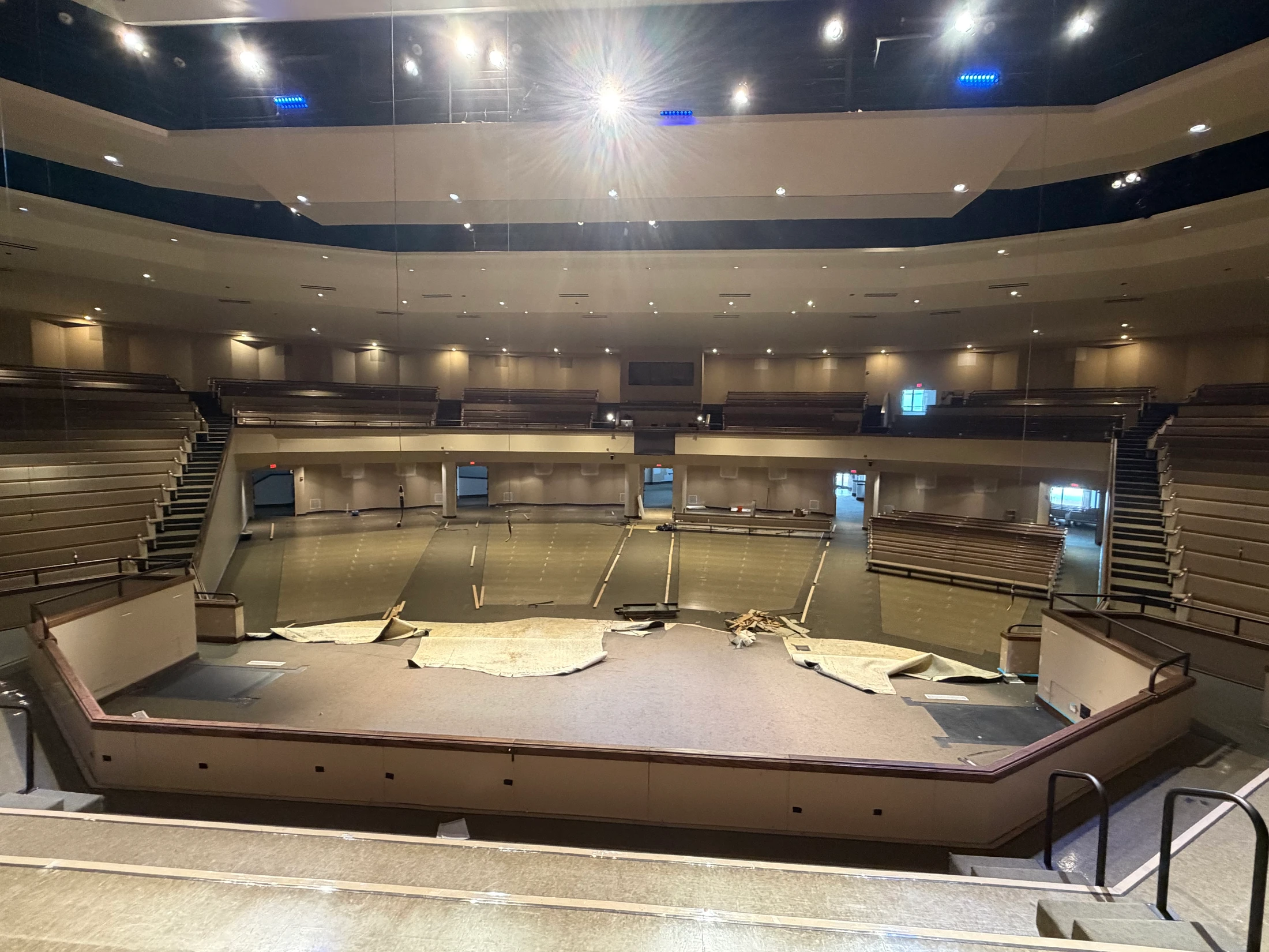 A wide-angle view of an empty auditorium with tiered seating during a renovation, with the carpeting on the floor and stage partially ripped up.