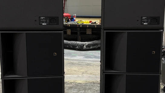 Two stacks of large black L-Acoustics speakers on pallets in an auditorium under construction, with scaffolding and a large screen in the background.