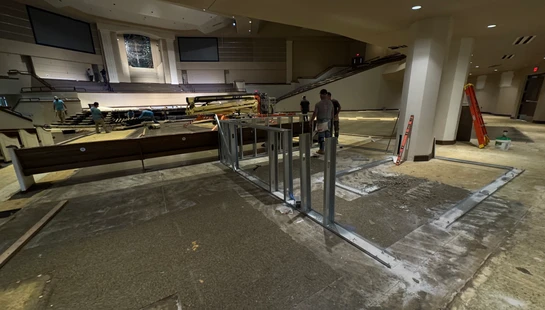 A wide view of a large church auditorium undergoing renovation. The floor is torn up, and workers are installing new metal wall framing in the foreground.
