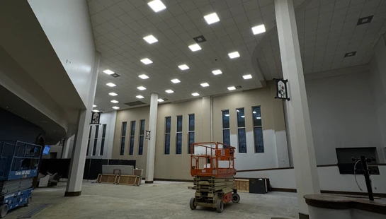 The interior of a large hall during renovation, with high ceilings, columns, construction lifts, and partially painted walls.