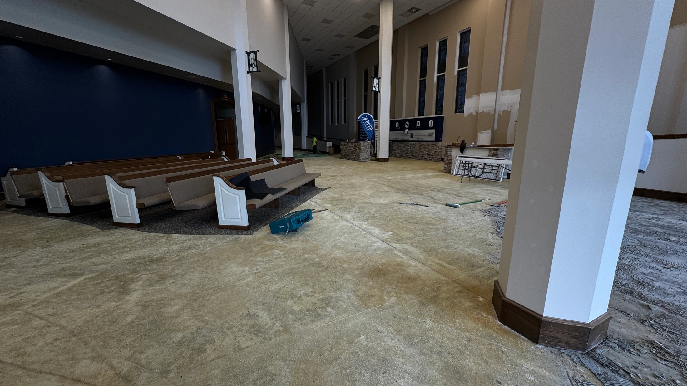 A wide view of a church interior during renovation. The old carpet is removed, exposing the concrete subfloor, with pews pushed to one side.